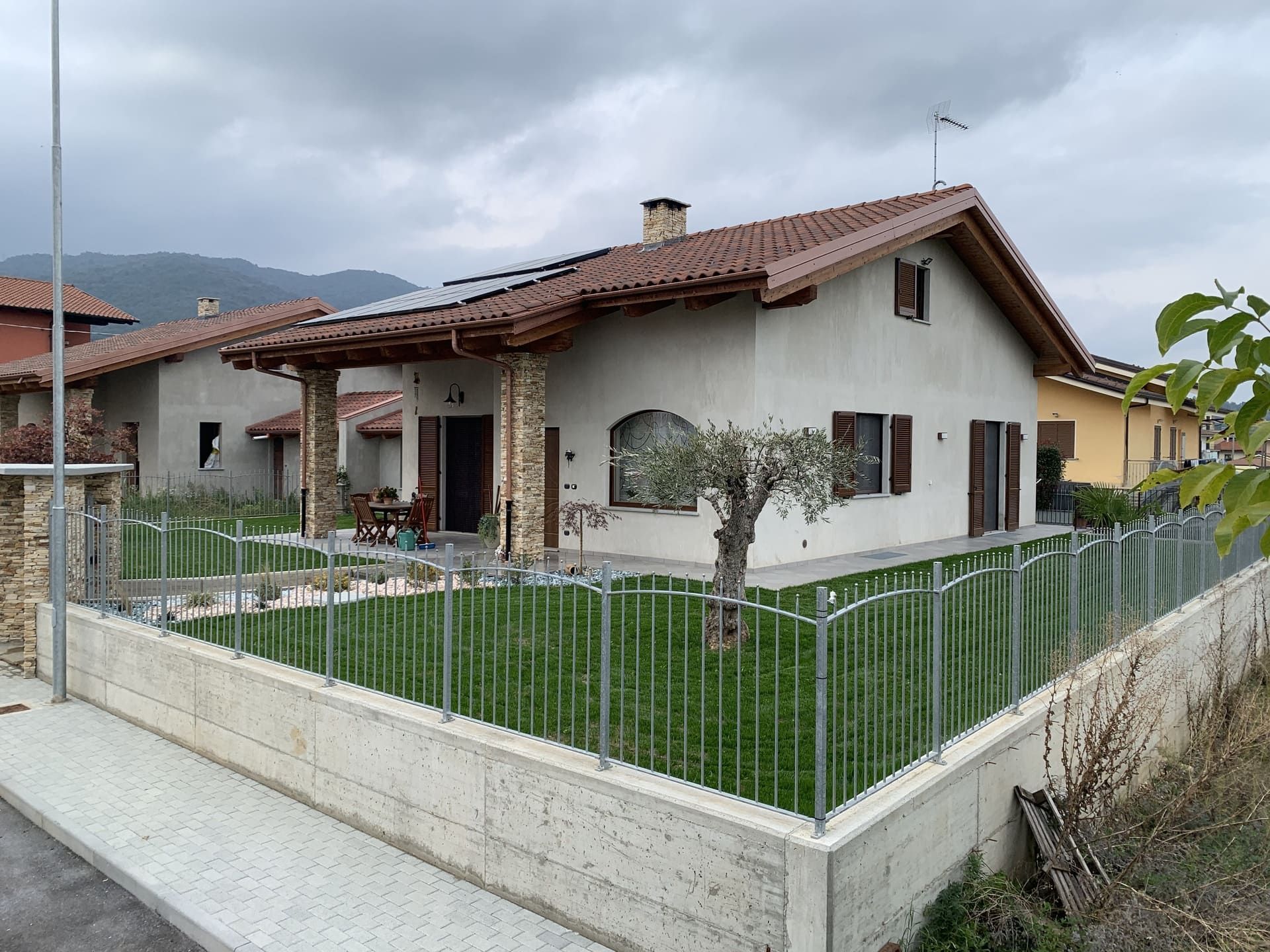 Una casa dai colori chiari con un tetto di tegole rosse, circondata da un prato verde e una recinzione metallica, il cielo è coperto.