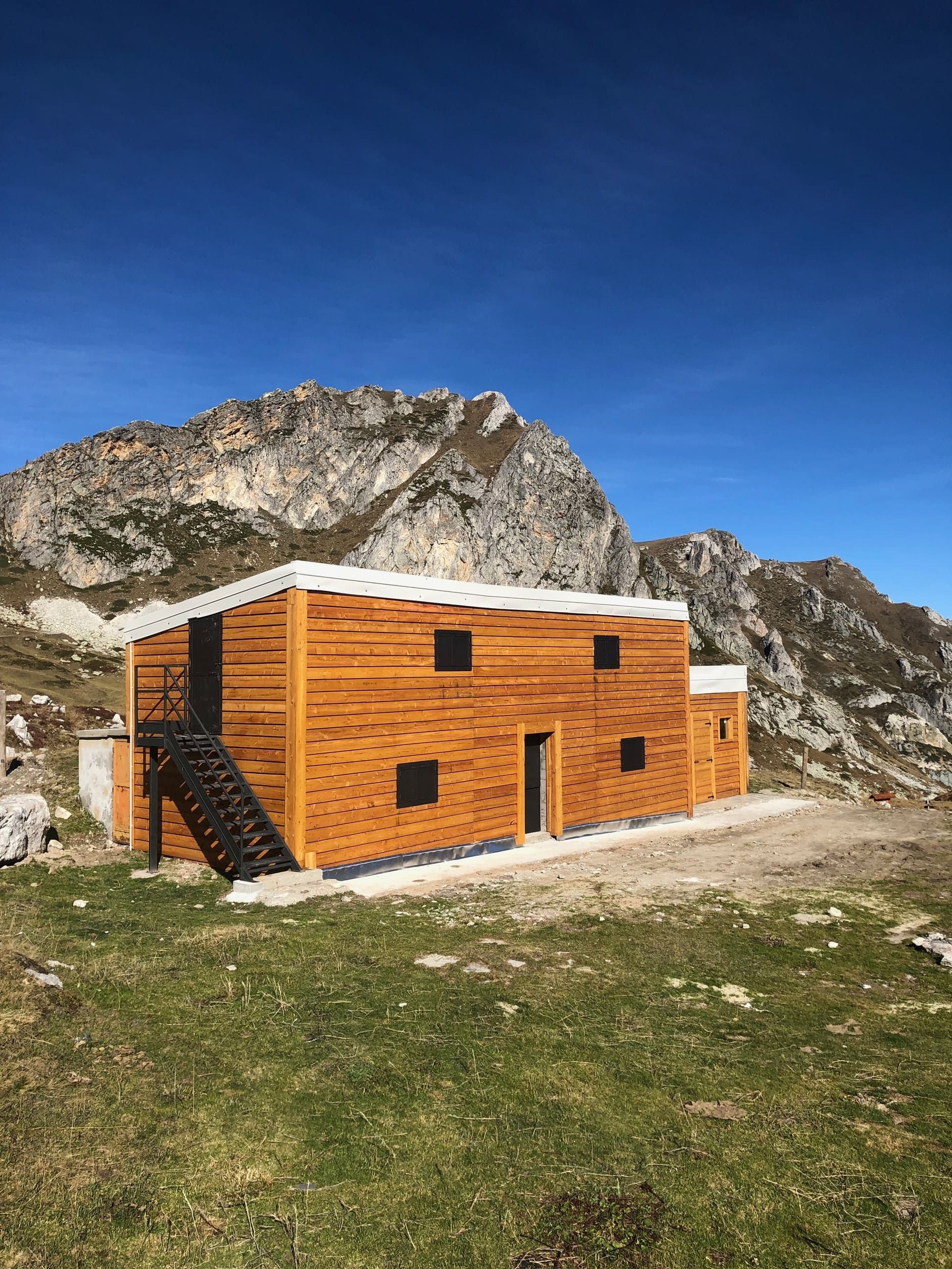 Baita di legno su un'area erbosa di fronte a una montagna rocciosa sotto un cielo azzurro.