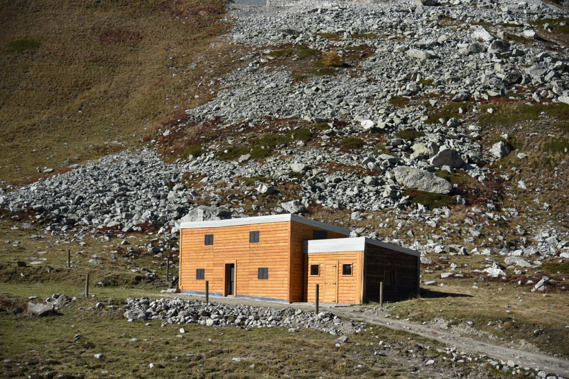 Edificio in legno in un paesaggio montano, caratterizzato da un ampio pendio roccioso e un sentiero sterrato.