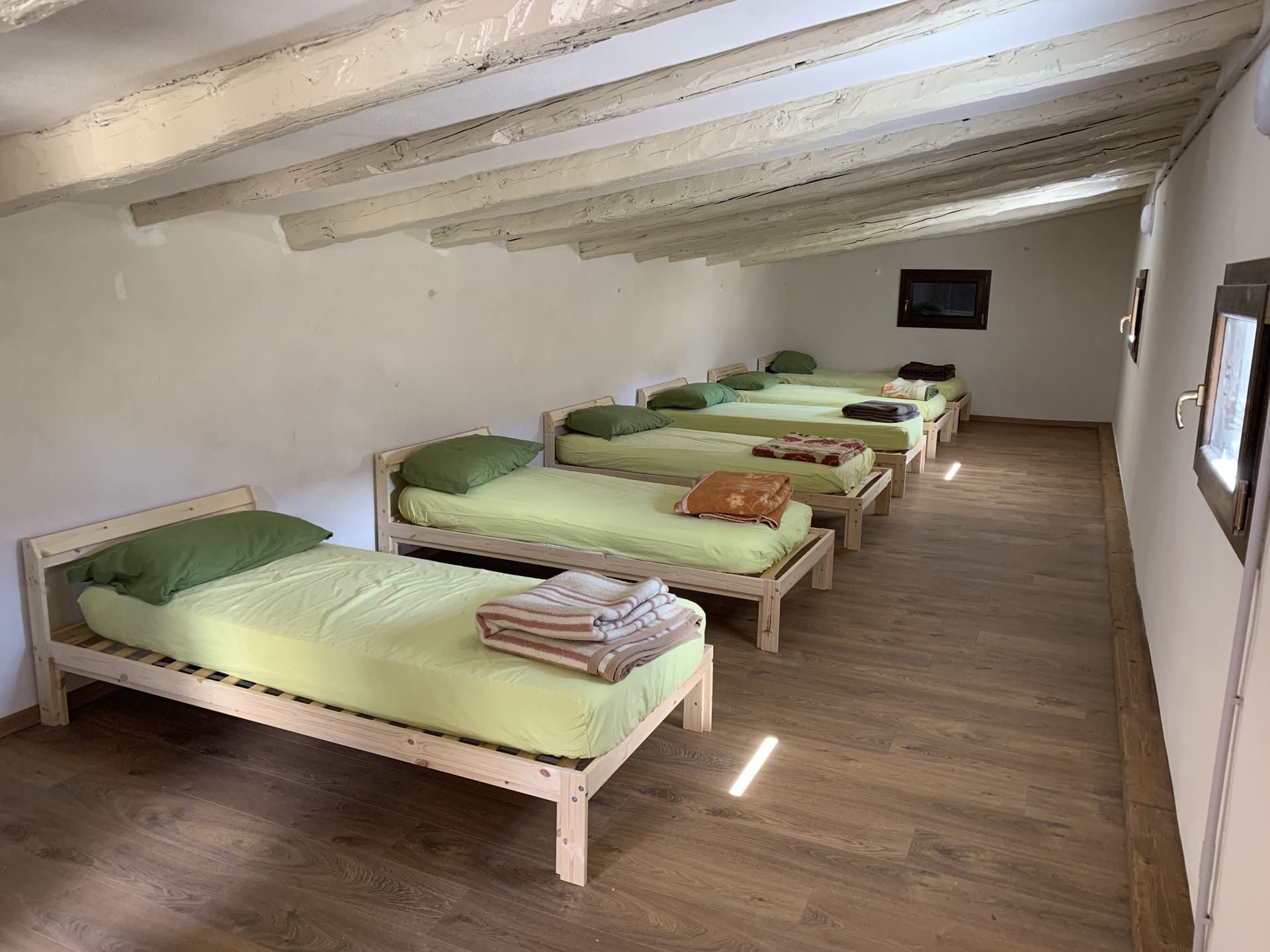 Letti in una stanza semplice, lenzuola verde chiaro, infissi e pavimento in legno. Pareti e soffitto bianchi, luce del sole che filtra dalle finestre.