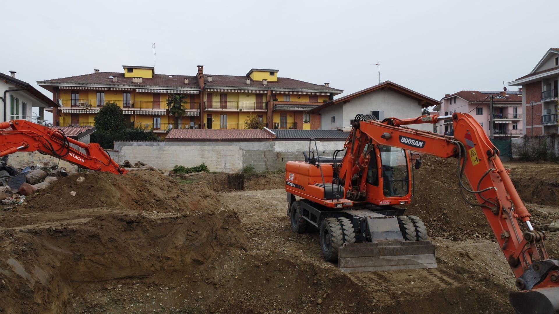 Callimaco - Centallo. Due escavatori arancioni scavano in un cantiere edile, con edifici sullo sfondo.