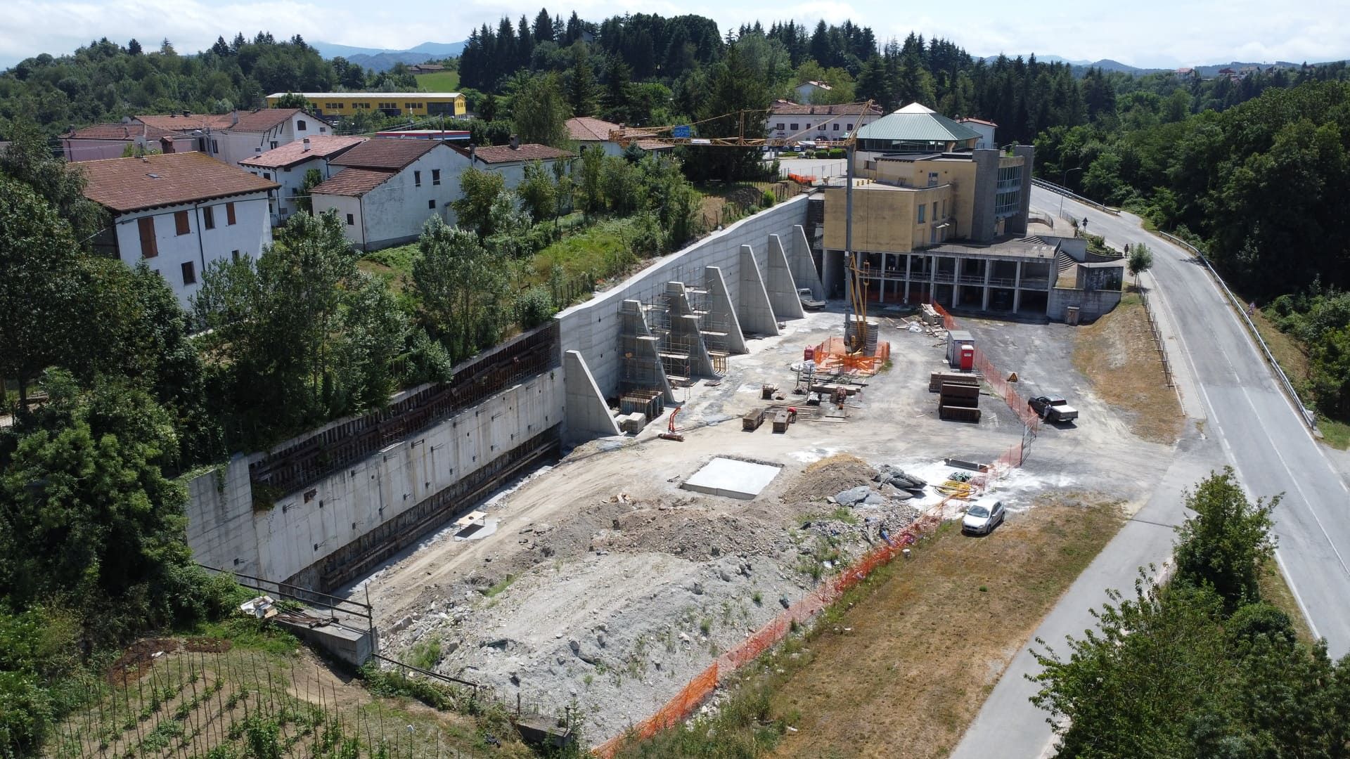 Cantiere con edifici e una strada, alberi circostanti, muri di contenimento.
