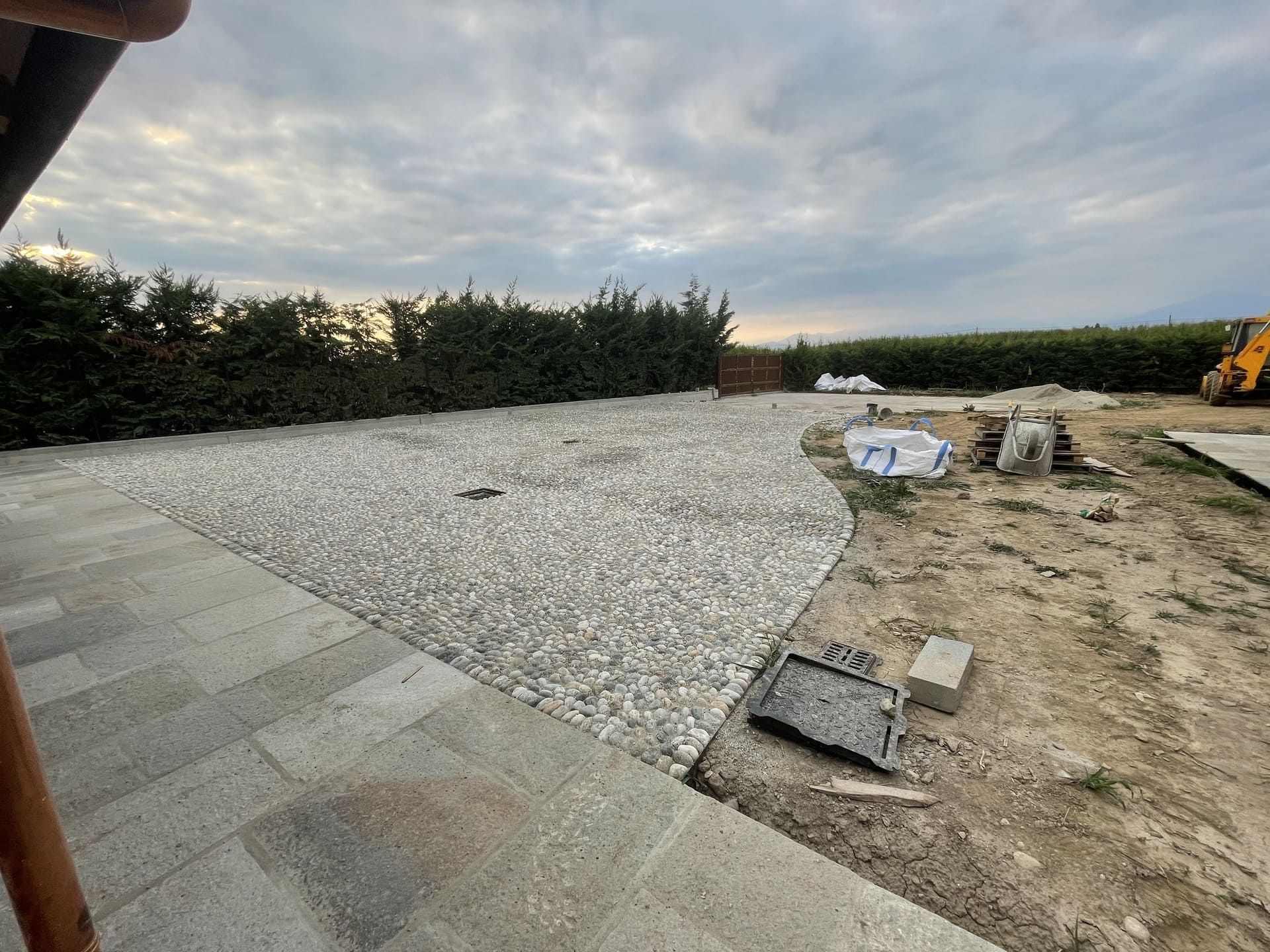 Cortile di ghiaia in costruzione con patio pavimentato, siepe verde e cielo nuvoloso.