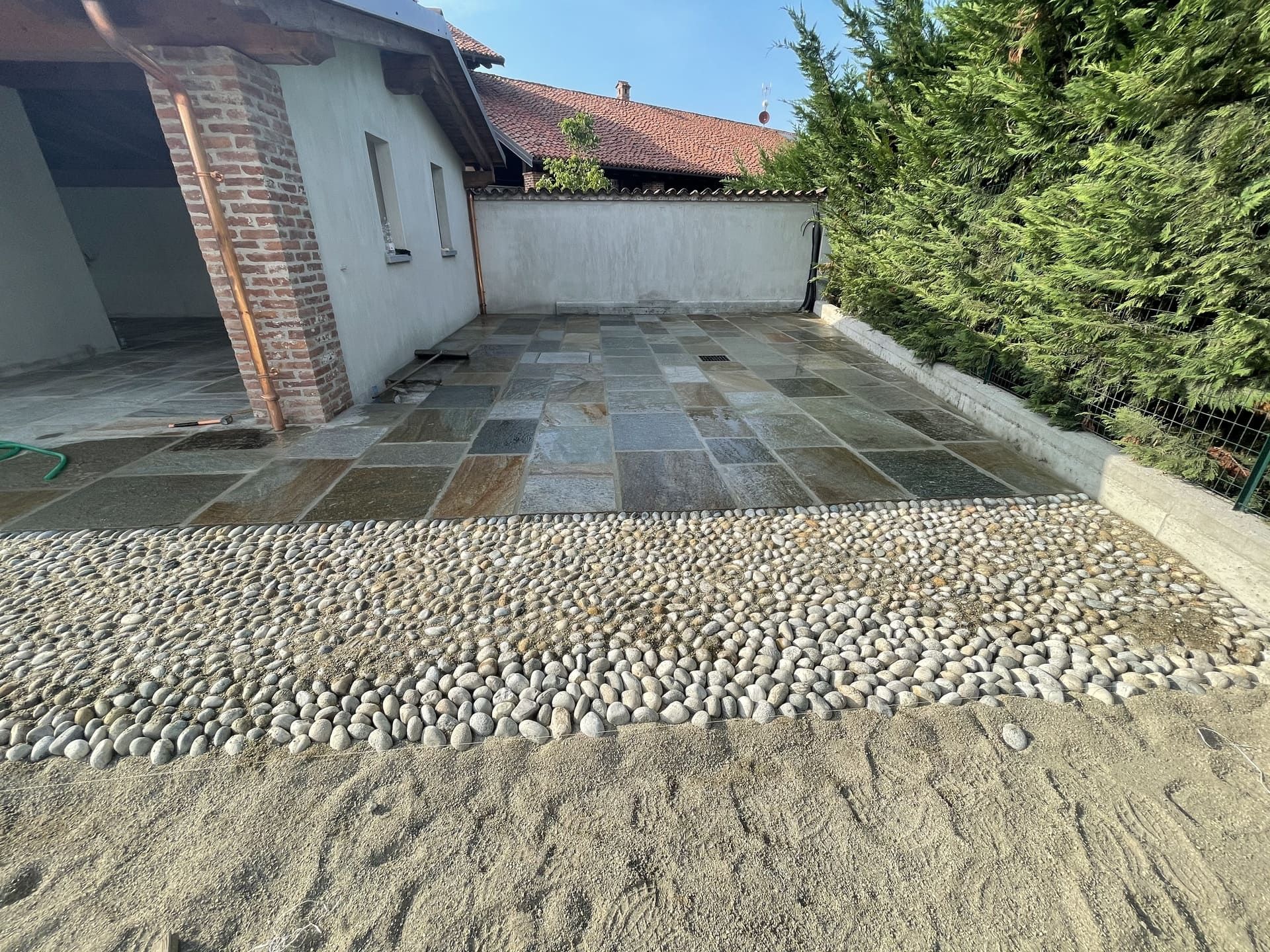 Patio in pietra con bordo in ciottoli, accanto a un edificio e al verde.