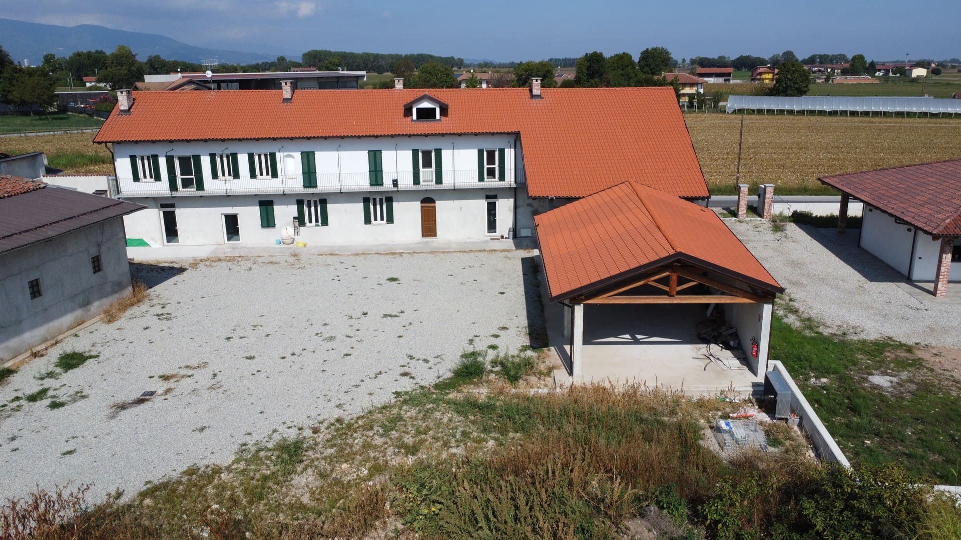 Un edificio bianco a due piani con tetto di tegole rosse, posto auto coperto e vialetto di ghiaia in un ambiente rurale.