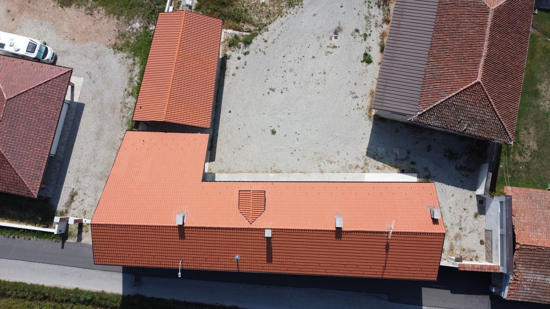 Vista aerea di edifici con tetti di tegole rosse disposti a L su un'area di ghiaia grigia.