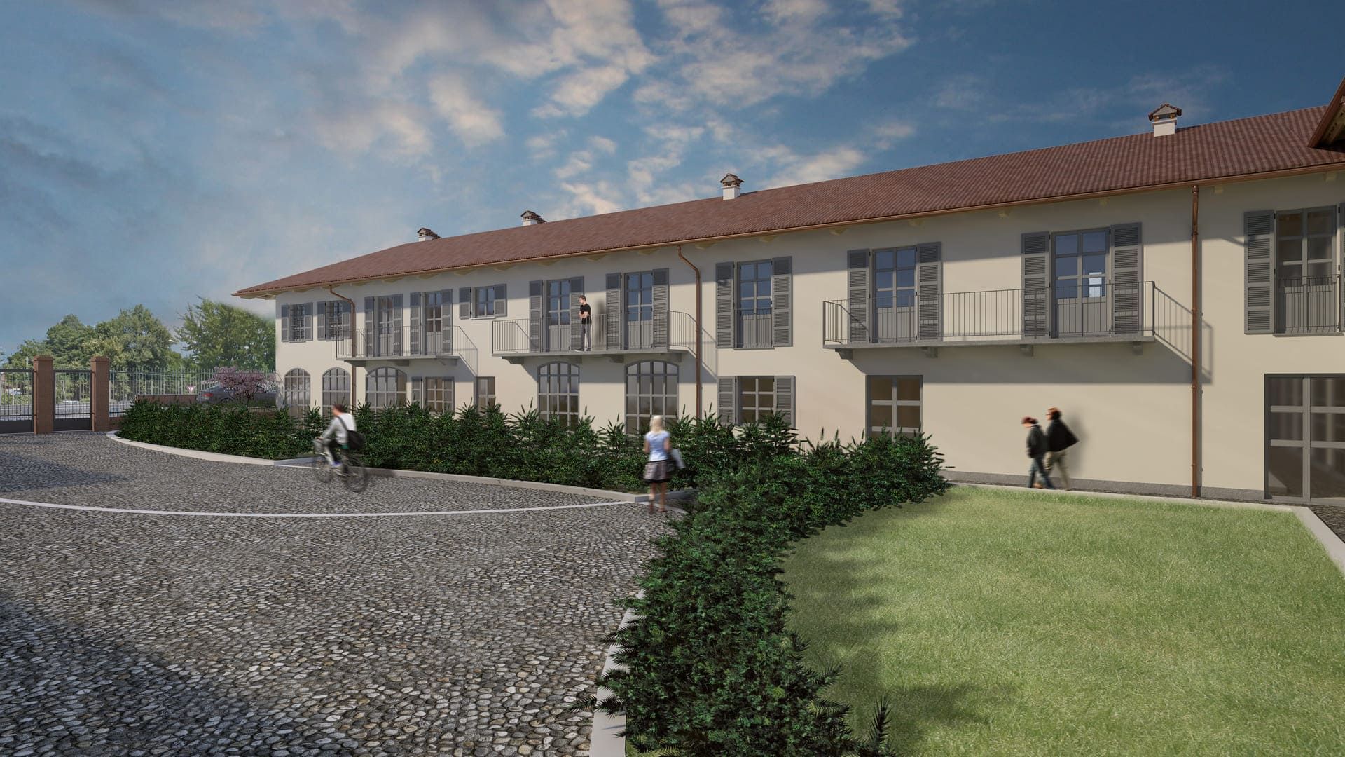 Un edificio dai colori chiari con balconi, tetto di tegole rosse e persone che camminano su un sentiero di pietra e su un prato verde sotto un cielo azzurro.