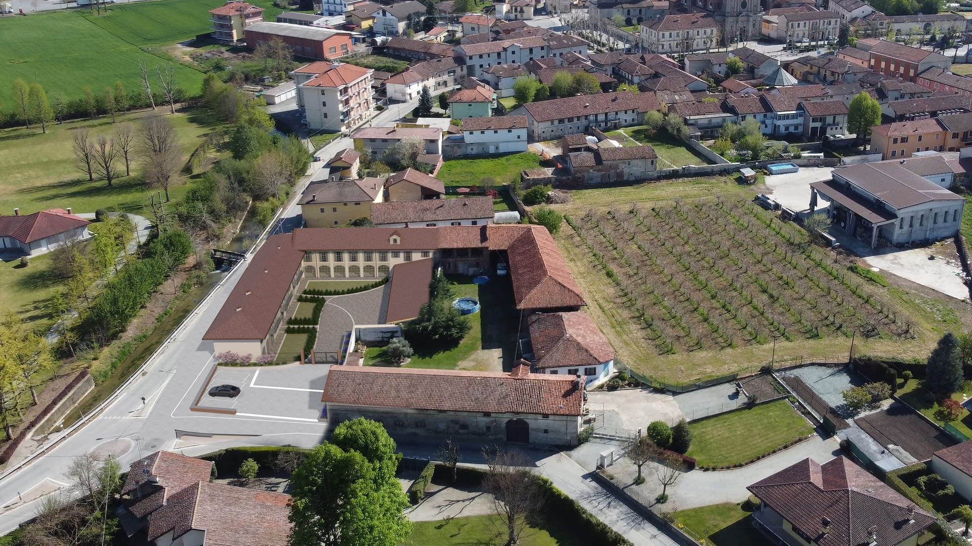 Vista aerea di un villaggio con edifici dai tetti rossi, strade e una grande struttura rettangolare al centro.