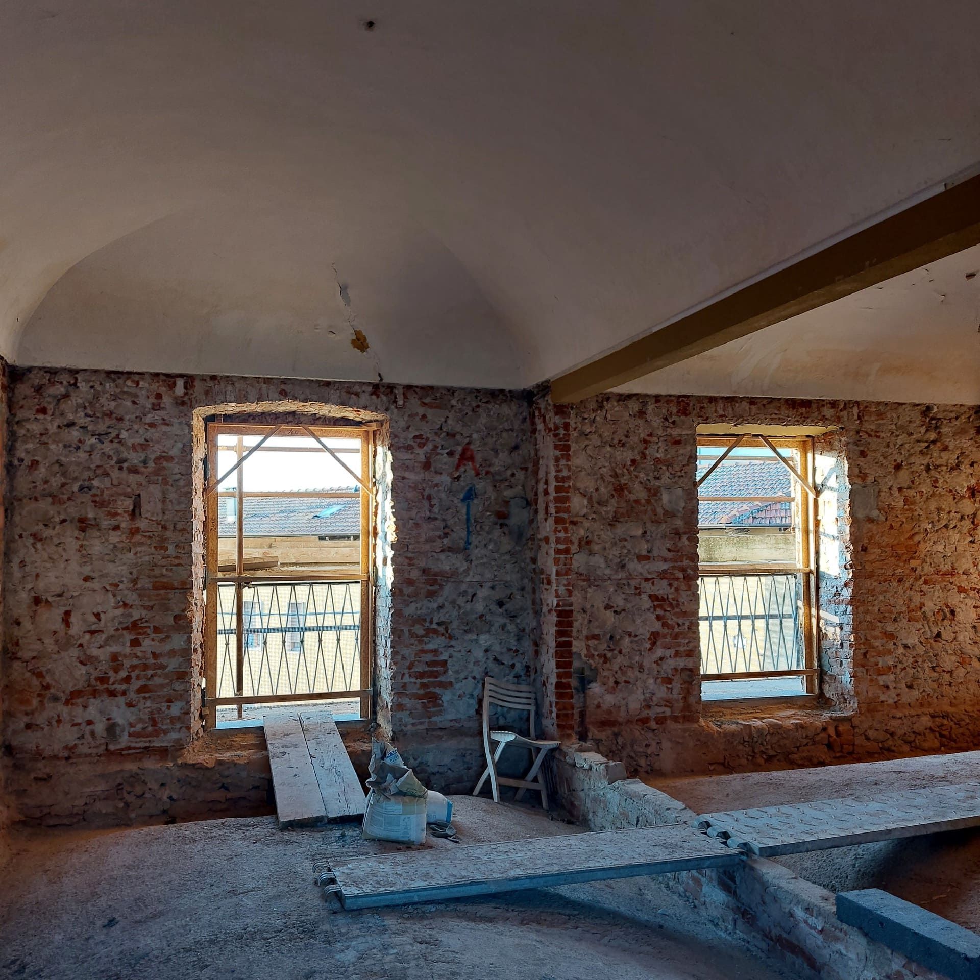 Vista interna di una stanza in costruzione con due finestre, pareti in mattoni a vista e soffitto curvo.