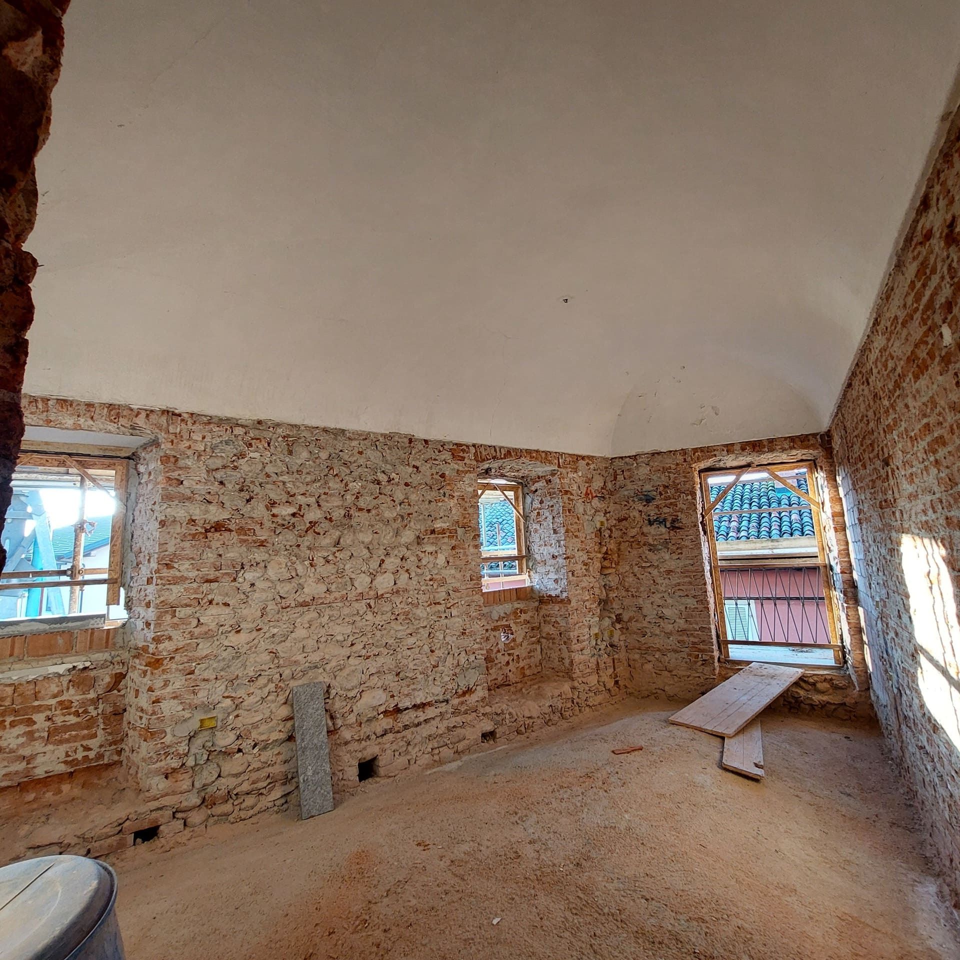 Interno di una stanza in fase di ristrutturazione: pareti con mattoni a vista, soffitto bianco, finestre e un'asse di legno.