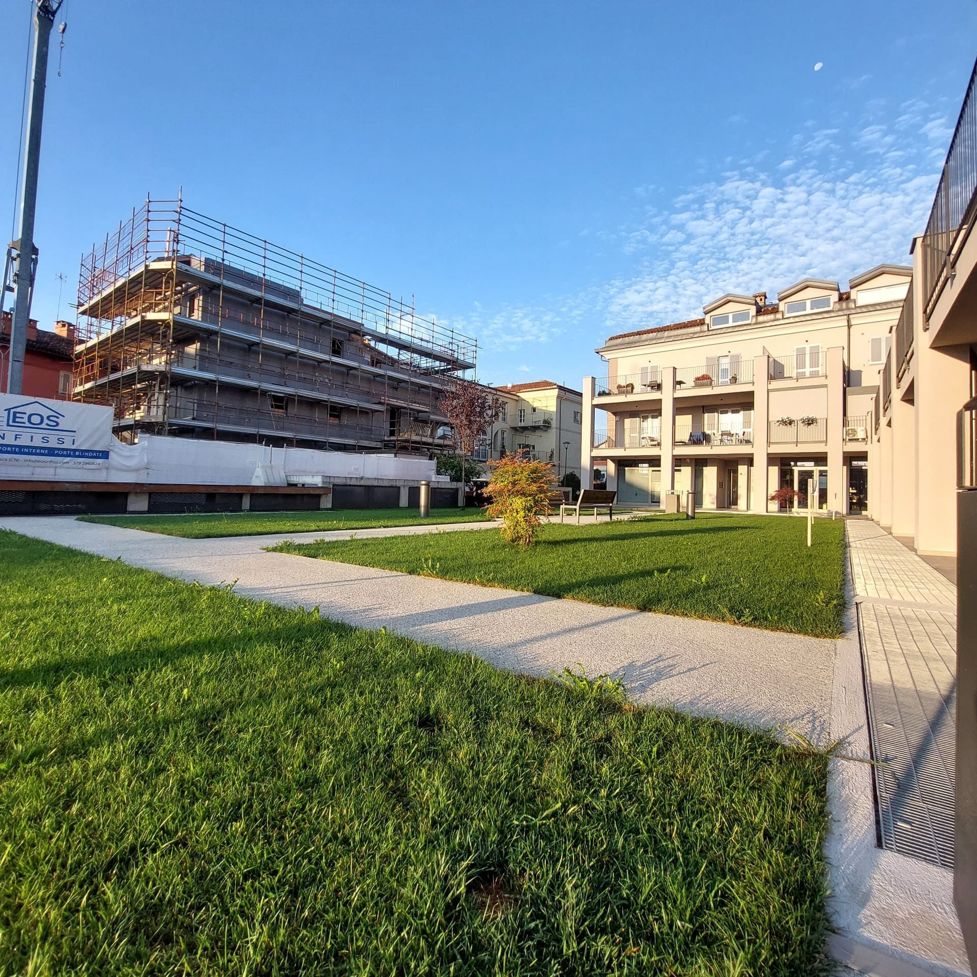 Prato verde con sentieri tra gli edifici, cantiere sullo sfondo. Cielo azzurro brillante.