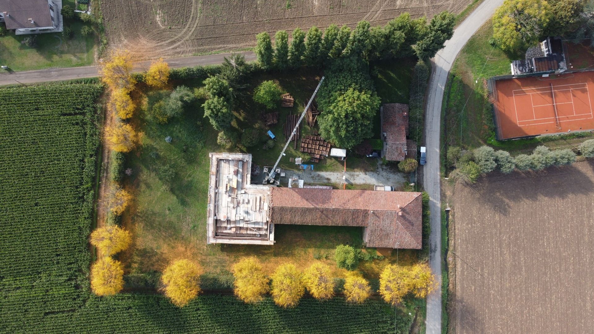 Vista aerea di un edificio con tetto in terracotta, circondato da alberi con foglie gialle, campi e un campo da tennis.
