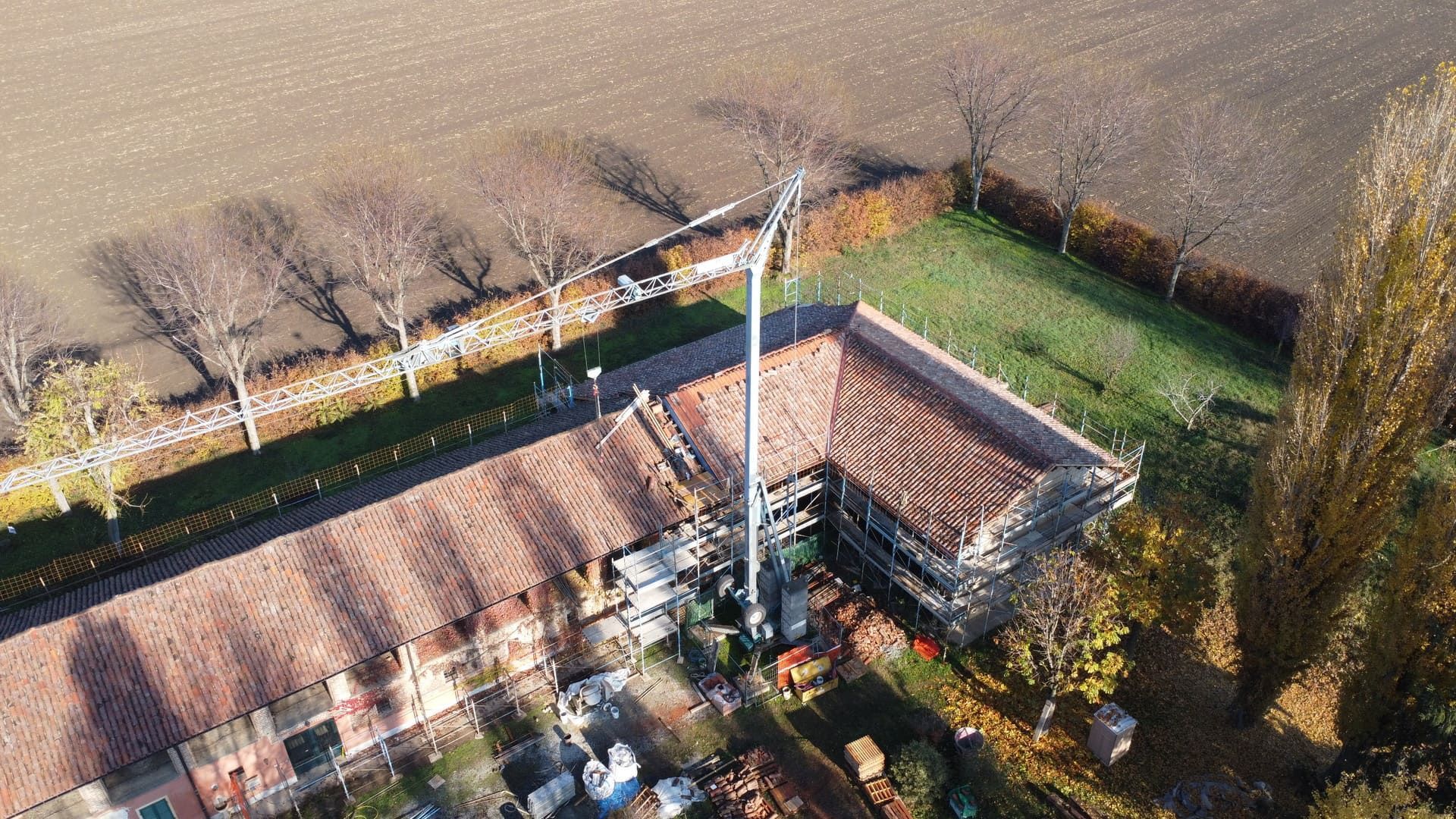 Vista aerea: Vecchio edificio con tetto in tegole rosse, impalcature, torre alta, campo marrone confinante e cortile verde.