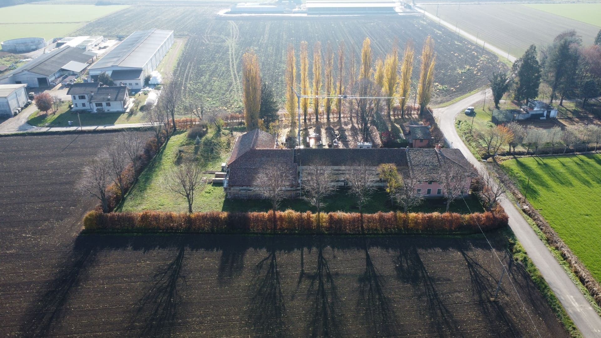 Vista aerea: edifici agricoli circondati da campi arati, alberi che proiettano lunghe ombre alla luce del sole.