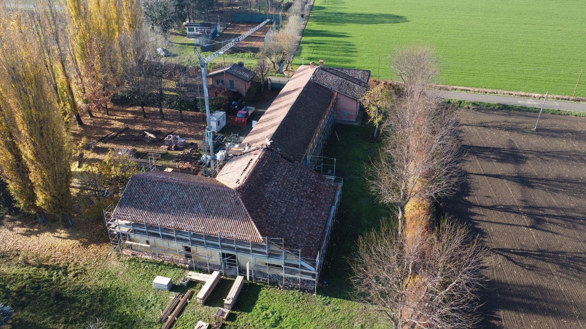 Vista aerea di un edificio rustico con tetto in tegole rosse, circondato da alberi e campi in autunno.