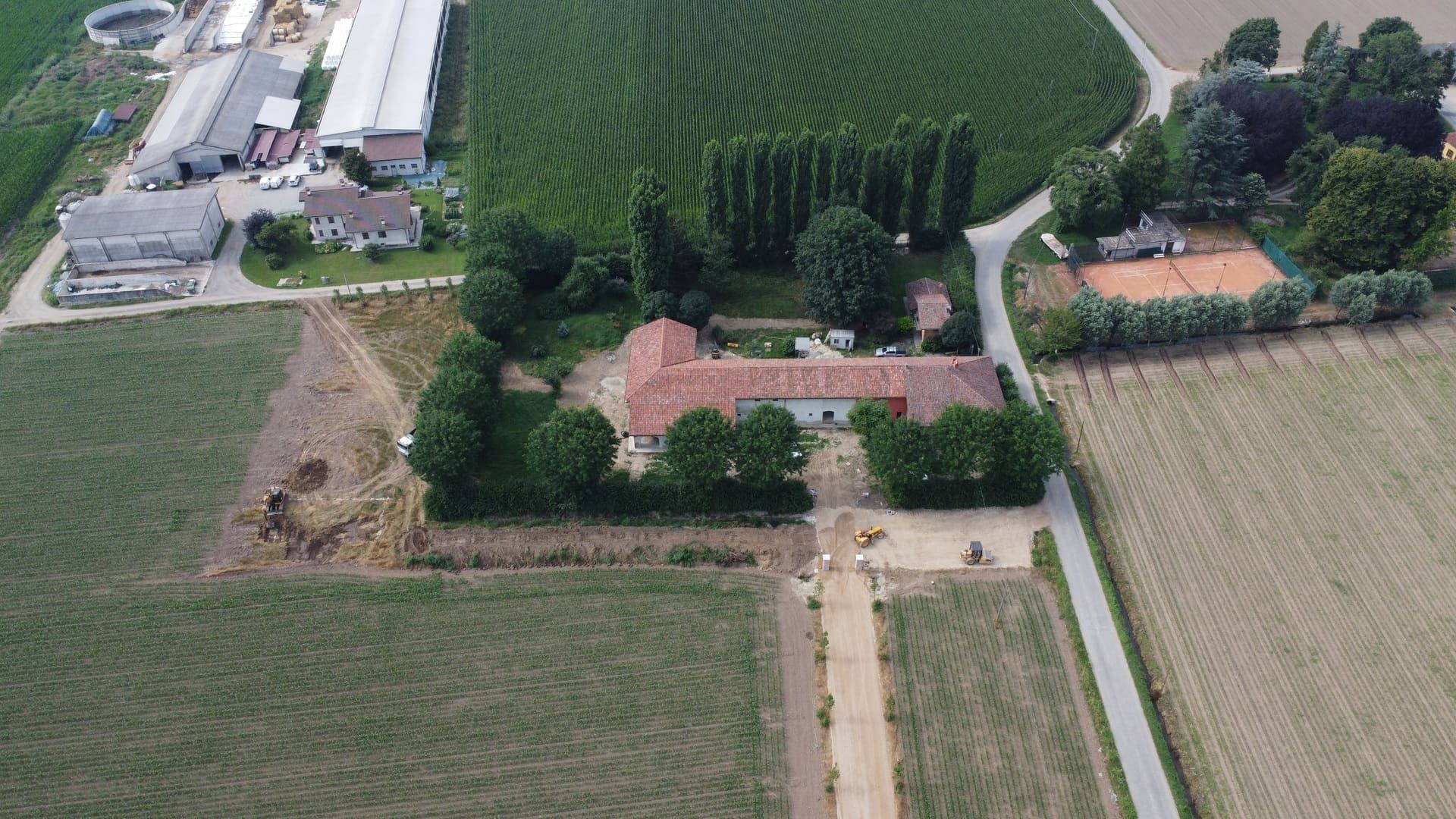 Vista aerea di una fattoria circondata da campi e alberi, con un campo da tennis sulla destra.