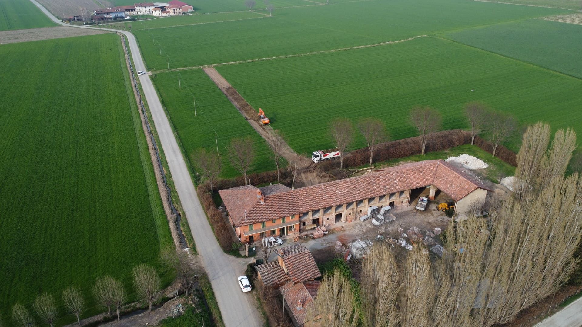Vista aerea di una fattoria con un lungo edificio in mattoni, una strada e campi. Sono presenti attrezzature edili.