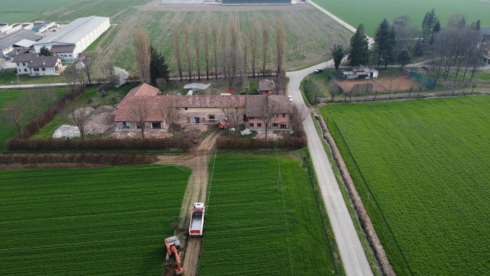 Un mezzo edile scava una trincea in un campo, vicino a un edificio agricolo. Campi verdi, alberi e una strada.