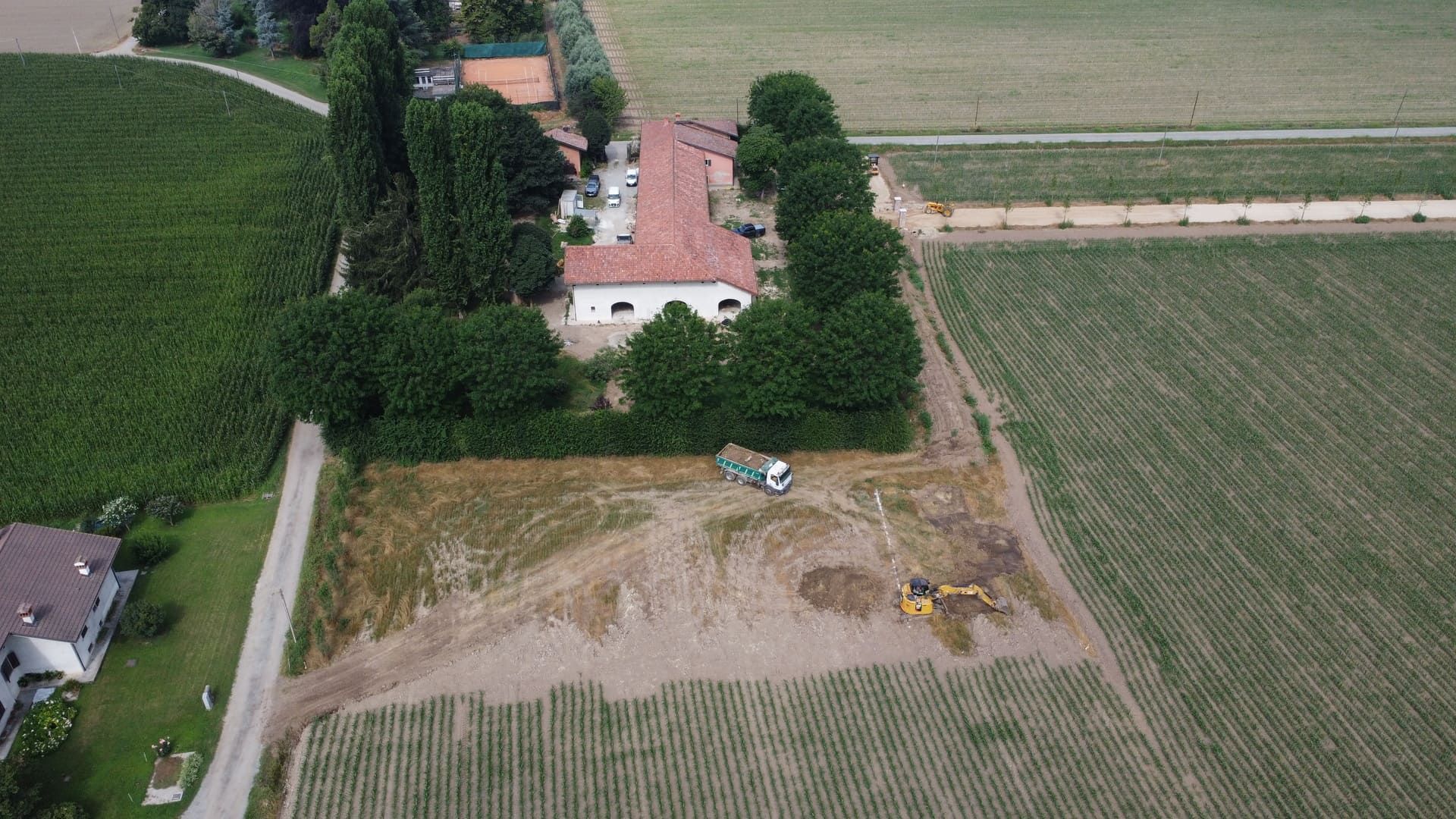 Vista aerea di un paesaggio rurale con una casa, alberi, campi e attrezzature edili.