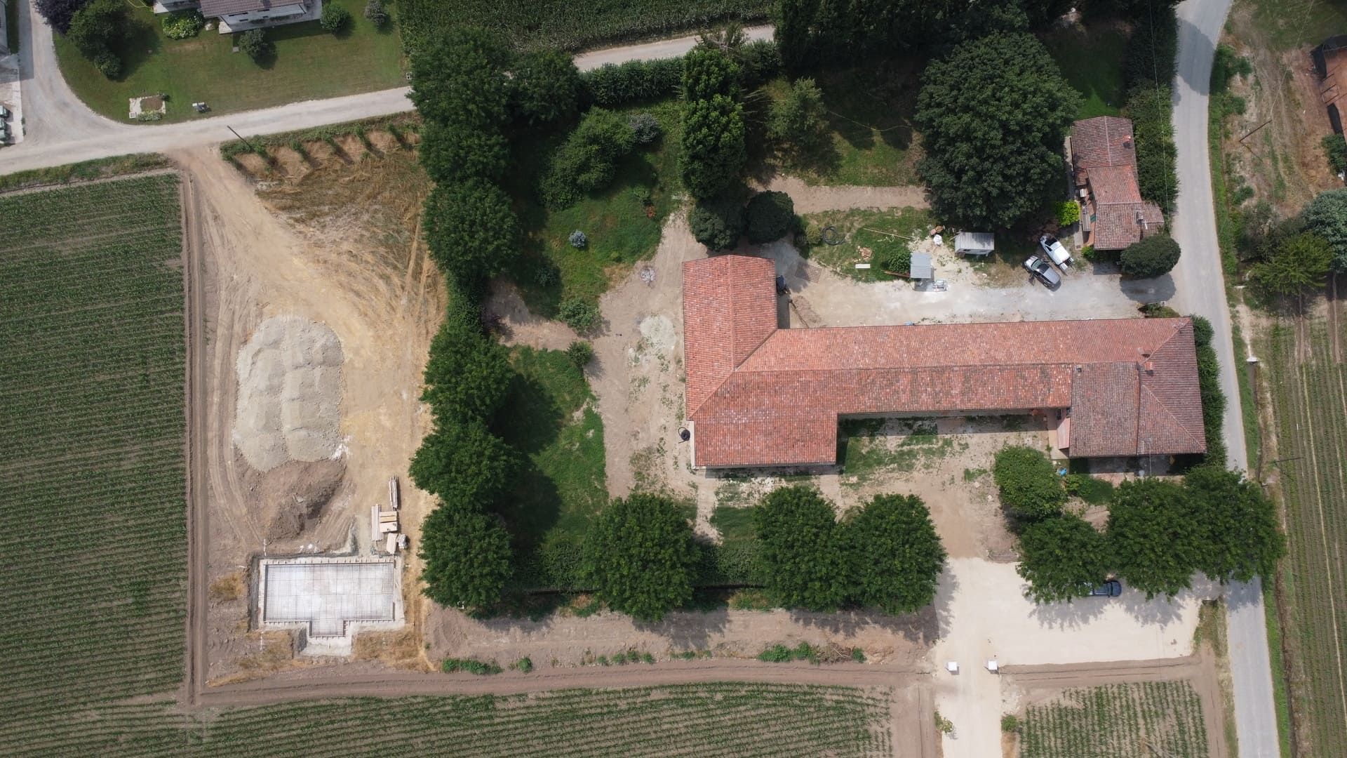 Vista aerea di un complesso di edifici con tetti rossi, circondato da alberi e campi. Lavori in corso nei pressi dell'edificio.