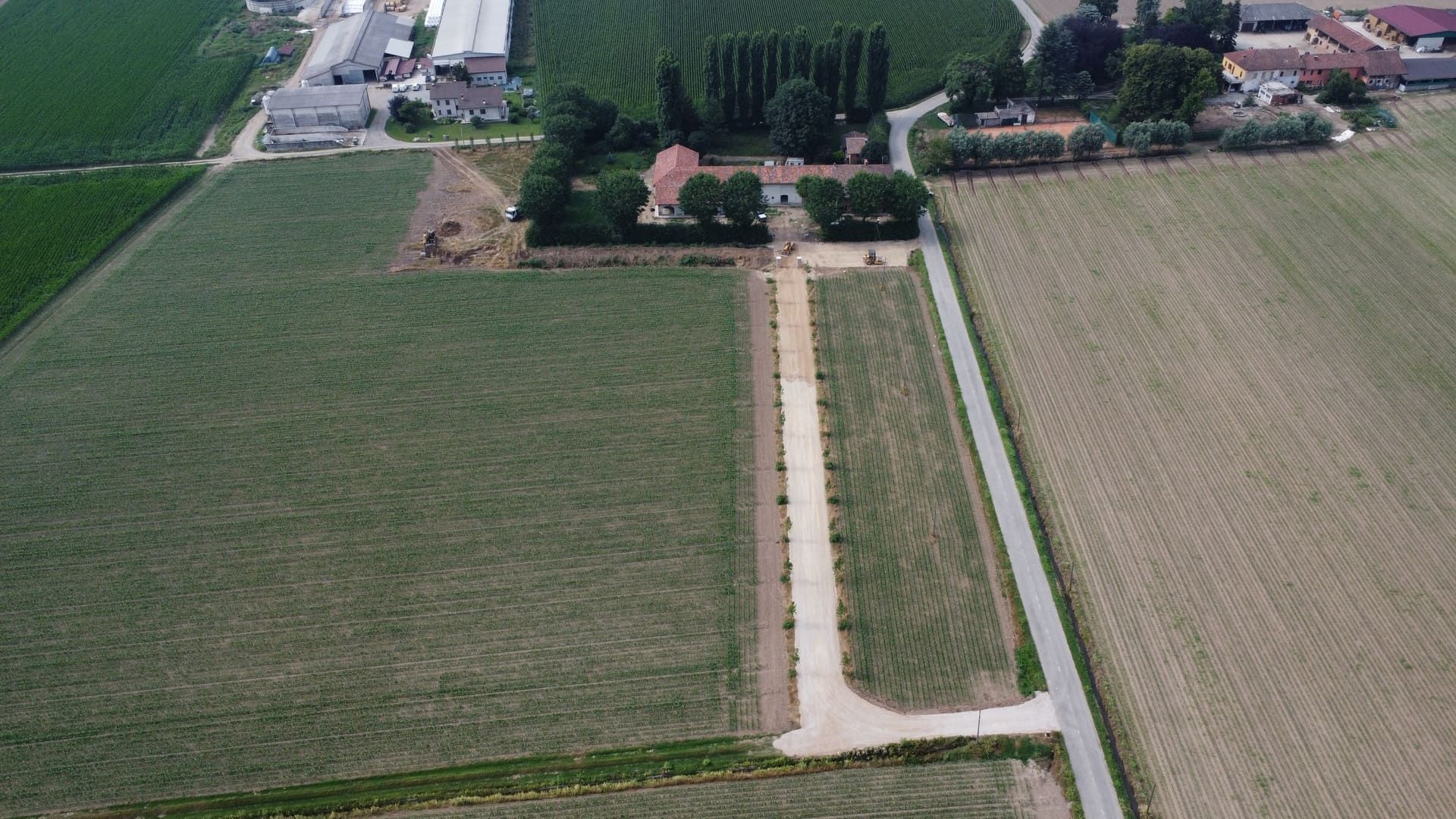 Vista aerea di un terreno agricolo con un sentiero sterrato che conduce a una casa, accanto a una strada asfaltata.
