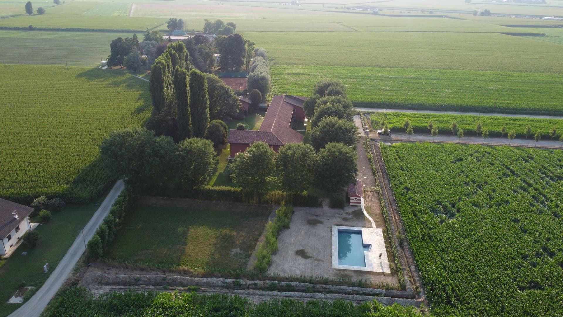 Vista aerea di una casa con piscina circondata da campi, alberi e una recinzione in un ambiente rurale.