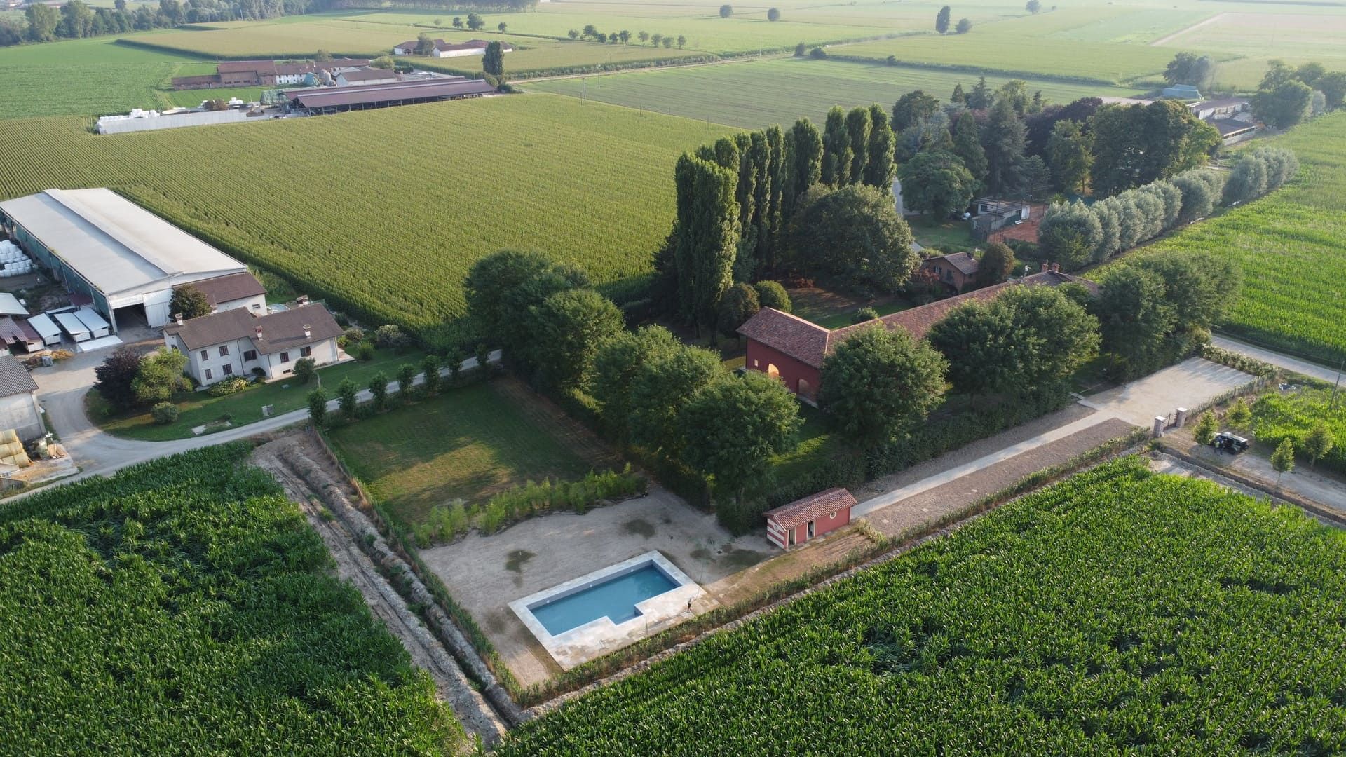 Vista aerea: edifici con tetti rossi, piscina, circondati da alberi e ampi campi verdi.