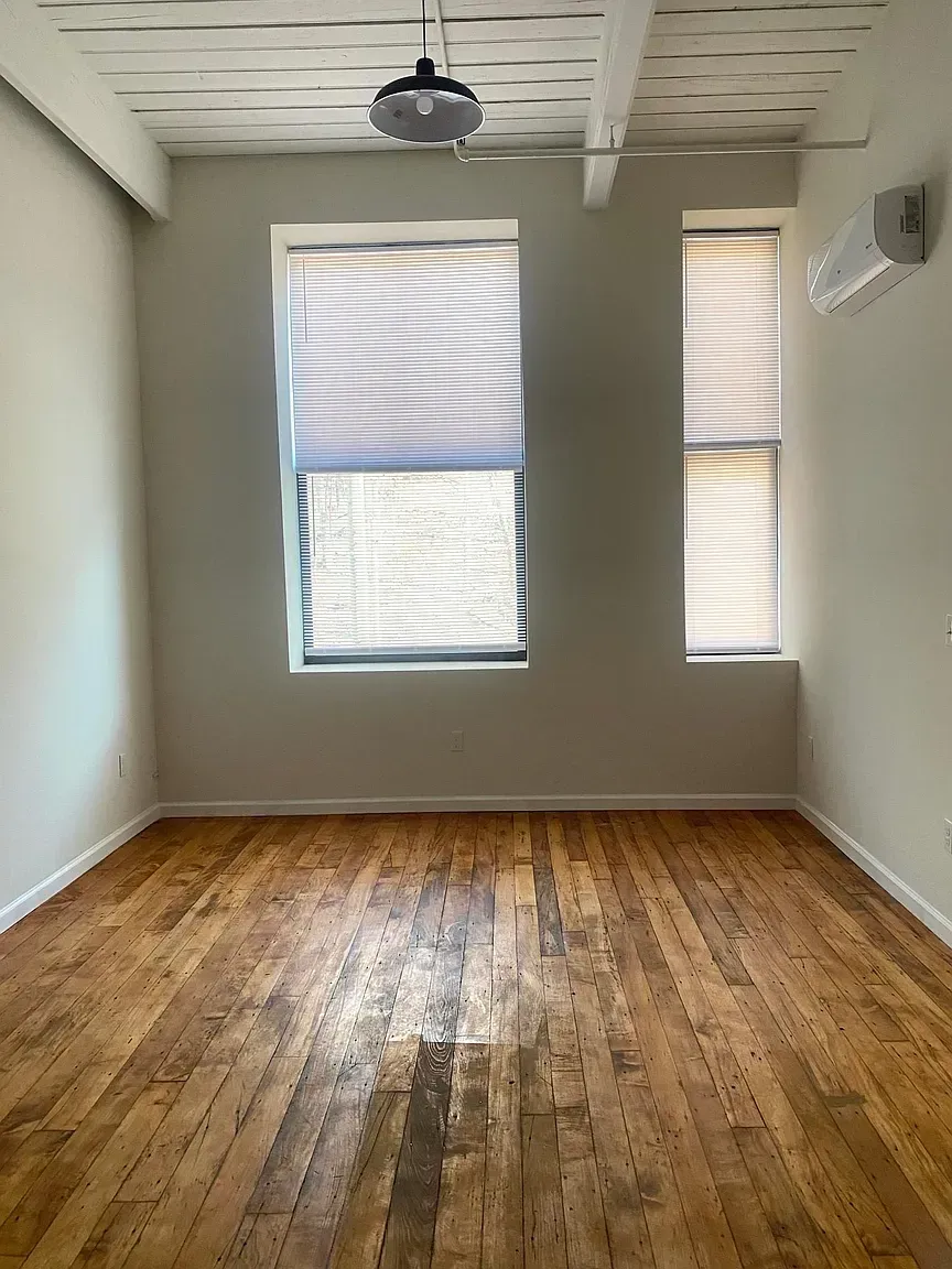 Empty room with hardwood floors, two windows with blinds, white walls, and a ceiling light.
