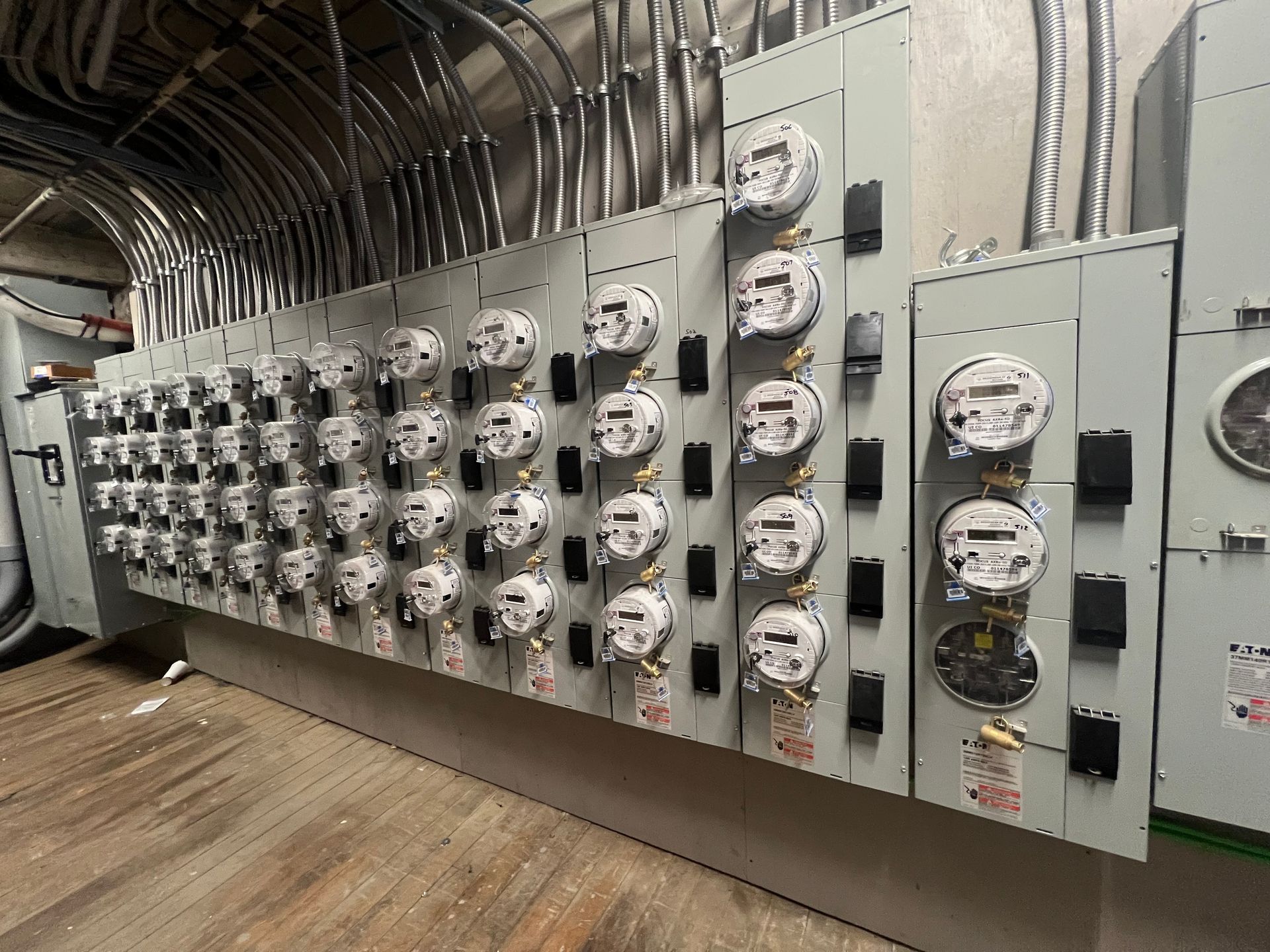 Electrical meters in a utility room, numerous digital displays on a gray wall, conduit overhead.
