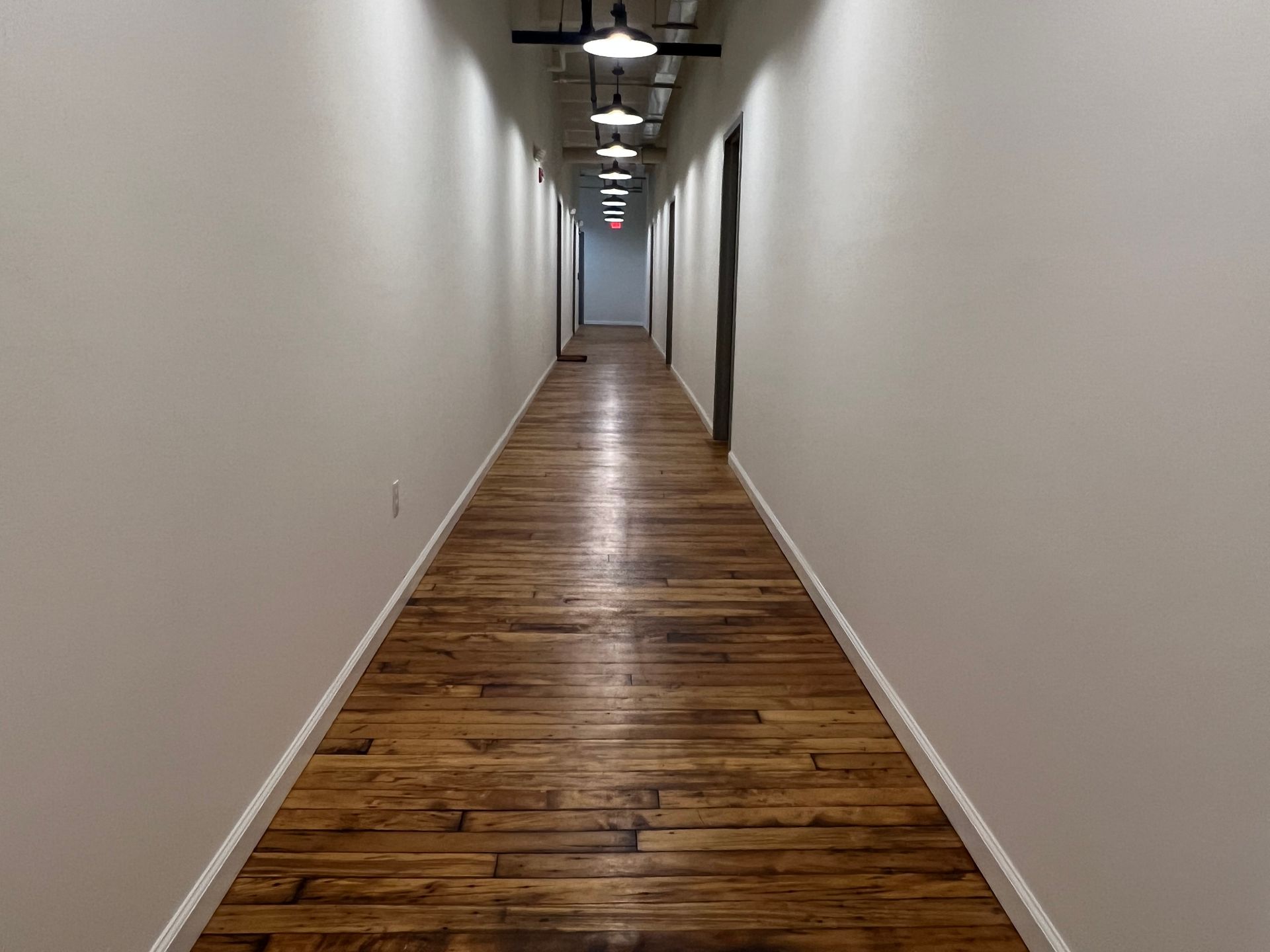 Long, narrow hallway with wooden floors, white walls, and overhead lighting.