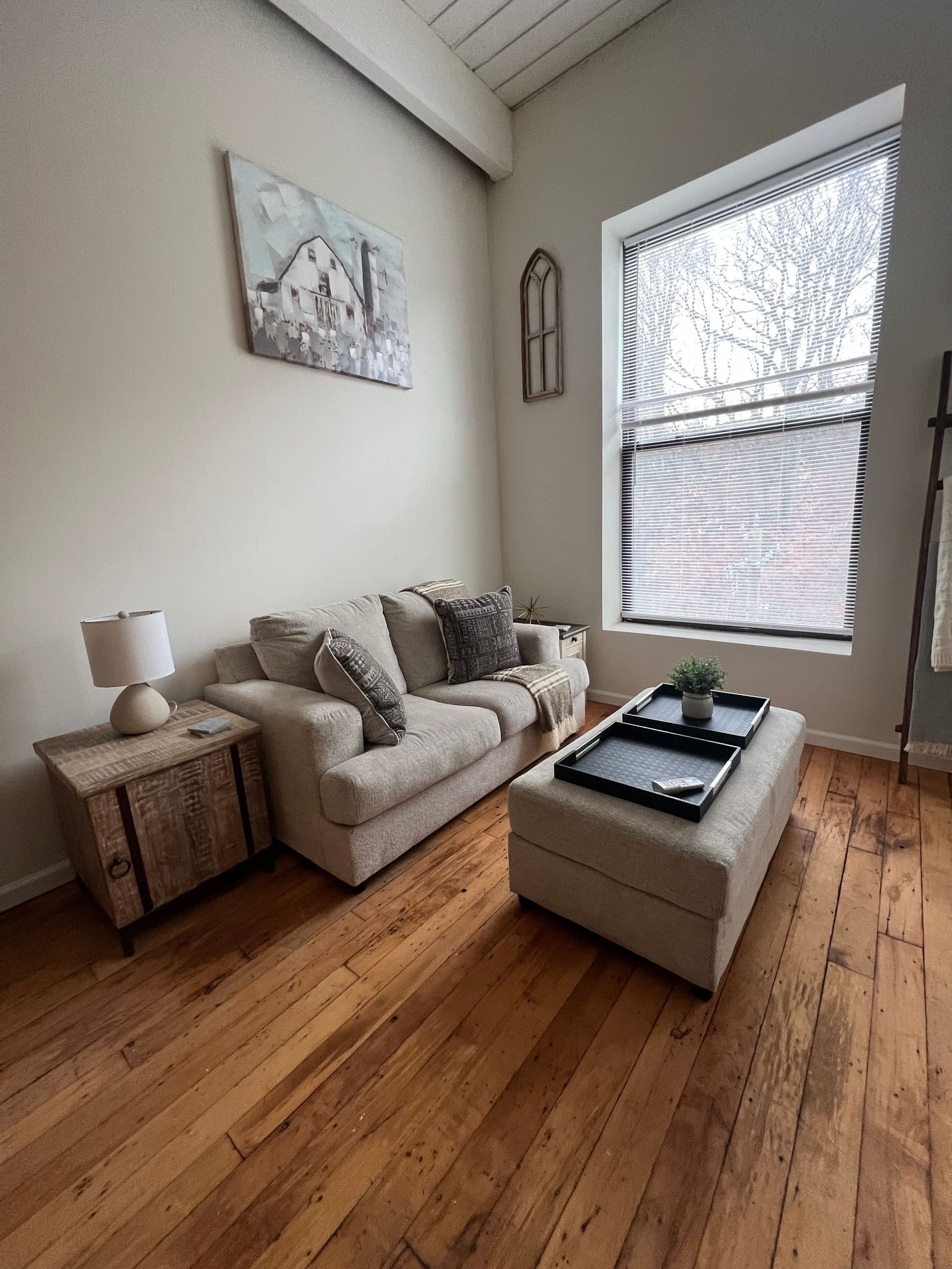 Cozy living room with a beige couch, ottoman, and wooden floor. A painting and window are visible.