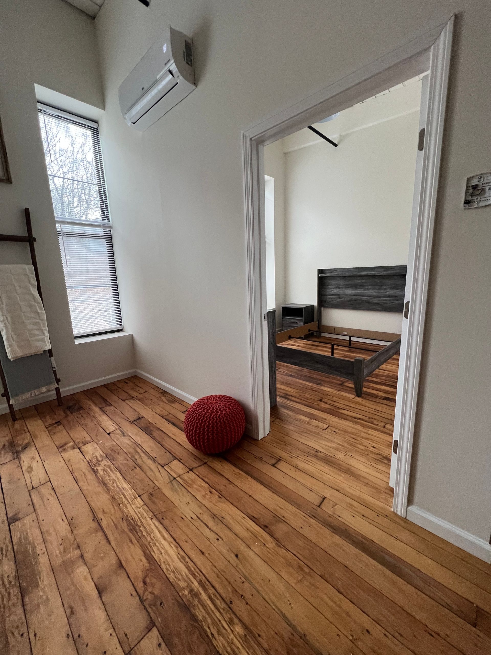 Bedroom with hardwood floors, doorway to a bed frame, red ottoman, window.