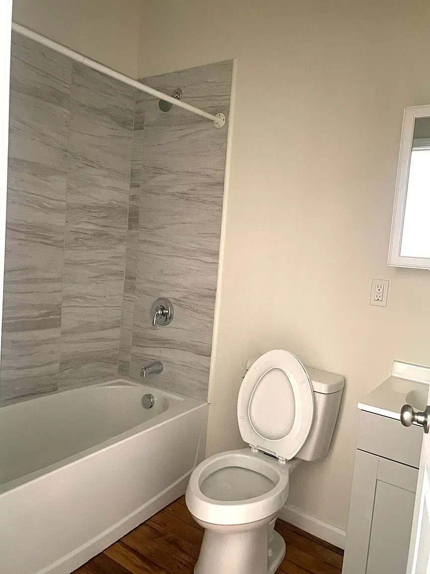 Bathroom with a white bathtub, gray-tiled shower, white toilet, and wooden floor.