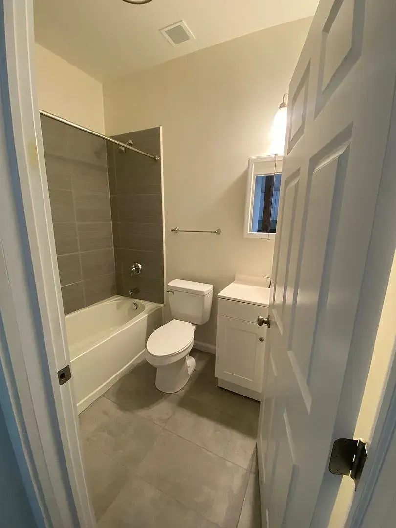 Bathroom with a white tub and toilet, gray tiled shower wall, and white vanity.