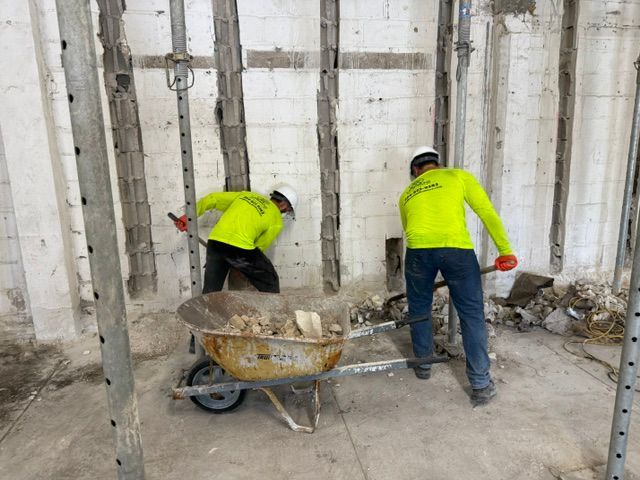 Two construction workers in neon yellow shirts shovel debris into a wheelbarrow inside a building with wall supports.