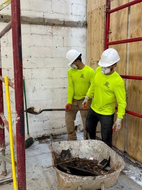 Two workers in high-visibility yellow long-sleeve shirts and white hard hats clearing debris into a wheelbarrow.