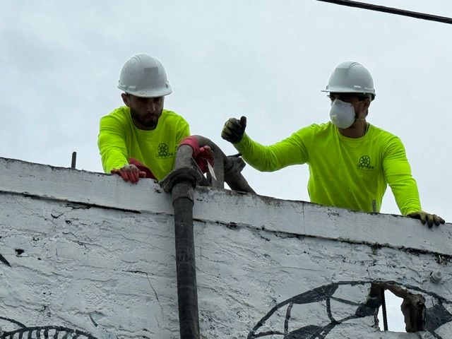 Two construction workers in white hard hats and neon yellow long-sleeve shirts hold a hose over a white exterior wall.