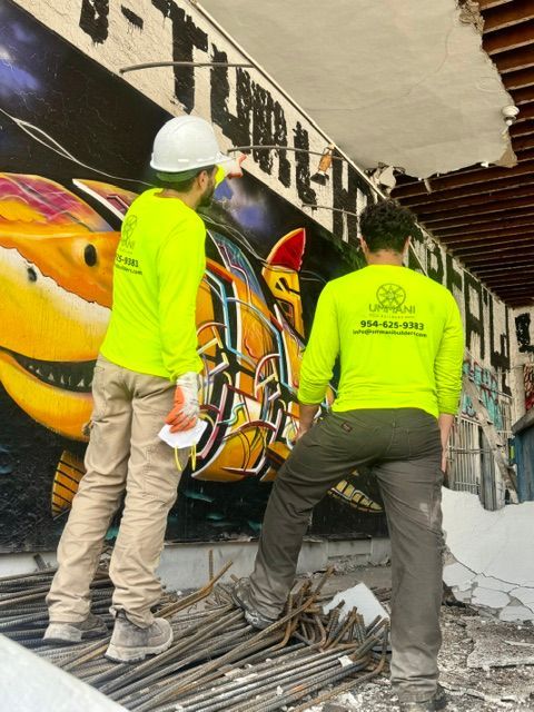 Two people in neon green long-sleeved shirts stand inside a construction site, looking at a colorful mural on a wall.