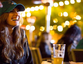 Woman smiling, wearing a baseball cap, holding a beer at an outdoor table, bokeh lights in background.