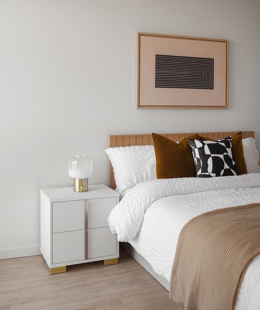 Bedroom with a white nightstand, bed with brown and black pillows, art, and a tan blanket.