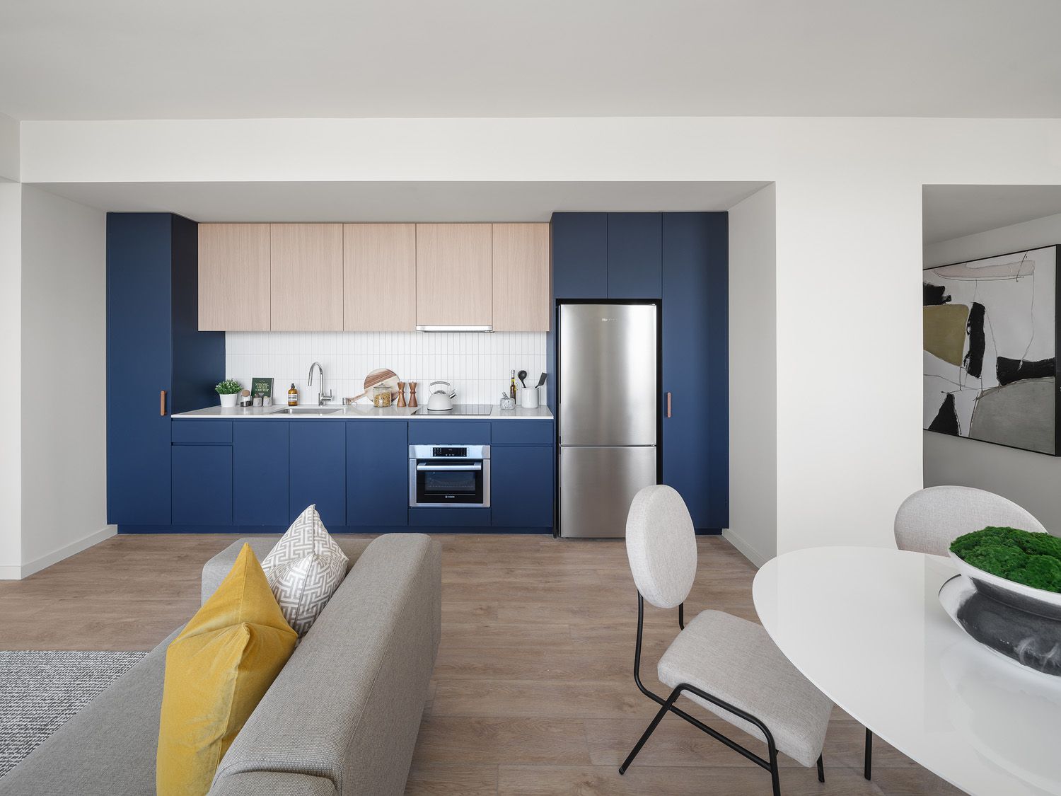 Modern kitchen with navy blue cabinets, light wood uppers, stainless steel appliances, and a living area with gray couch and dining table.