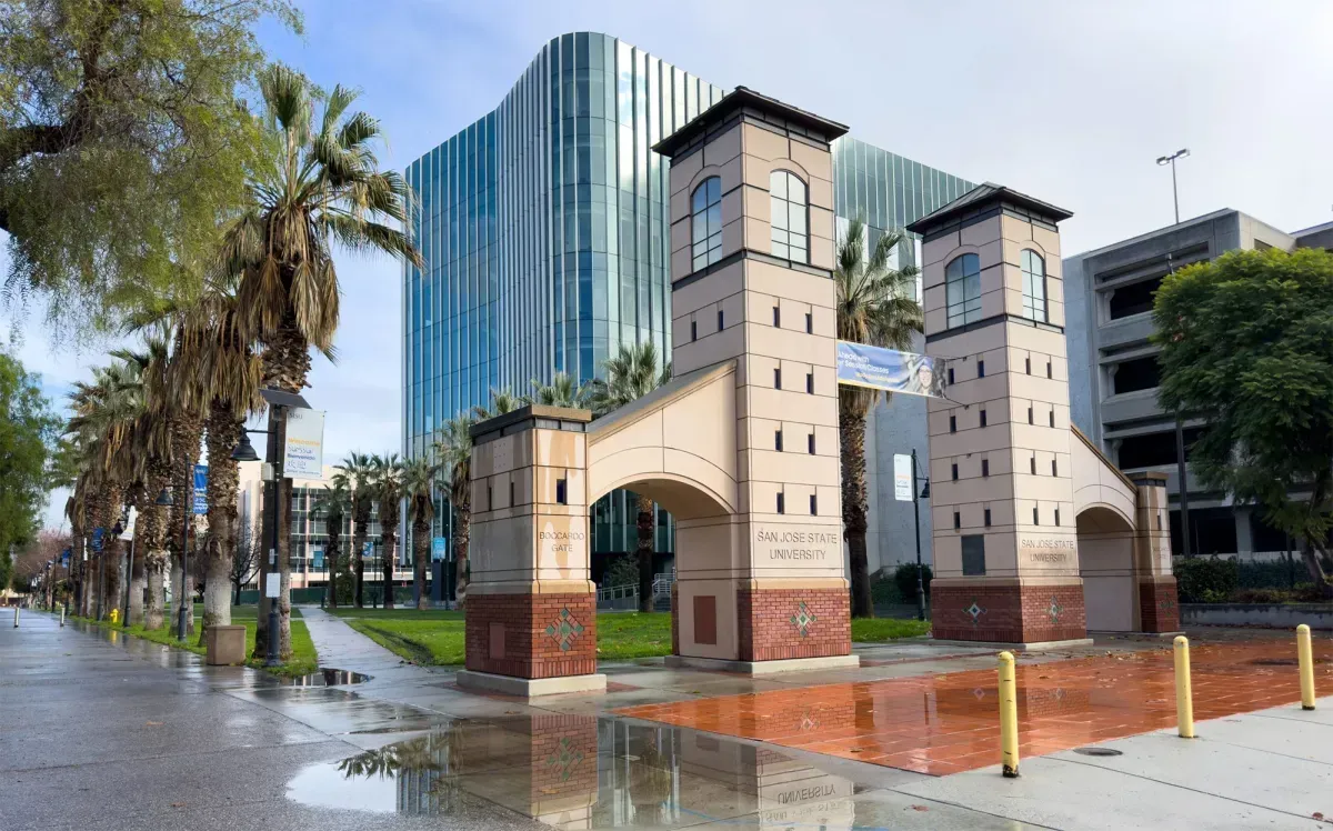 Brick archway with two towers in front of a modern glass building. Palm trees line the street.