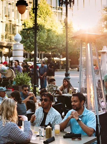 People seated at outdoor restaurant tables, illuminated by a sunlit heater. City backdrop.