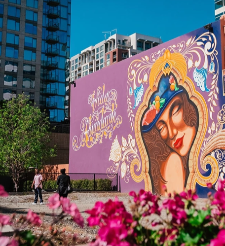 Mural with woman in ornate frame. Purple and gold details. Two people walk in front, pink flowers in foreground.