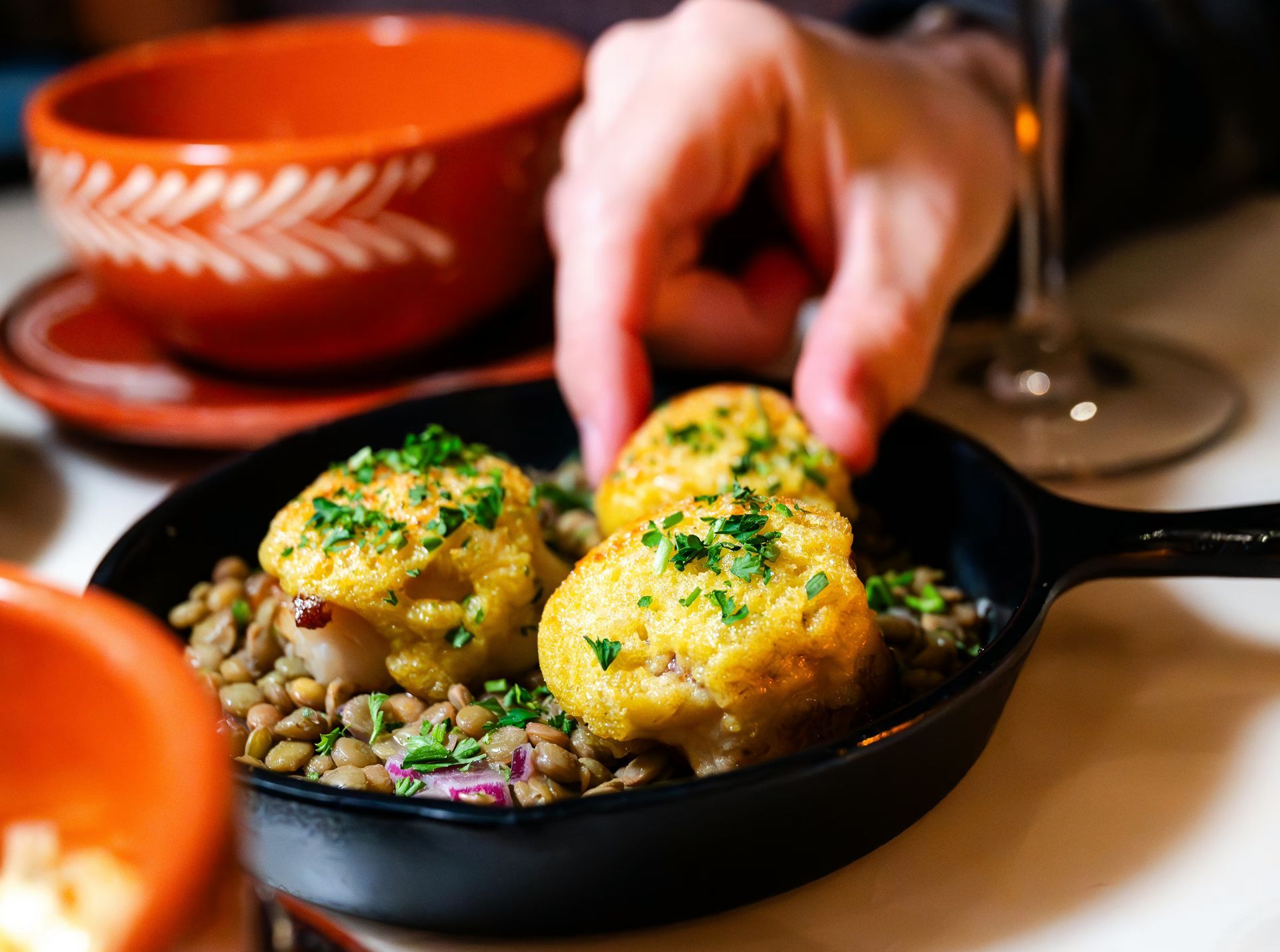 Hand reaching for three golden-topped appetizers on lentils in a cast iron dish; orange bowl and glass in background.