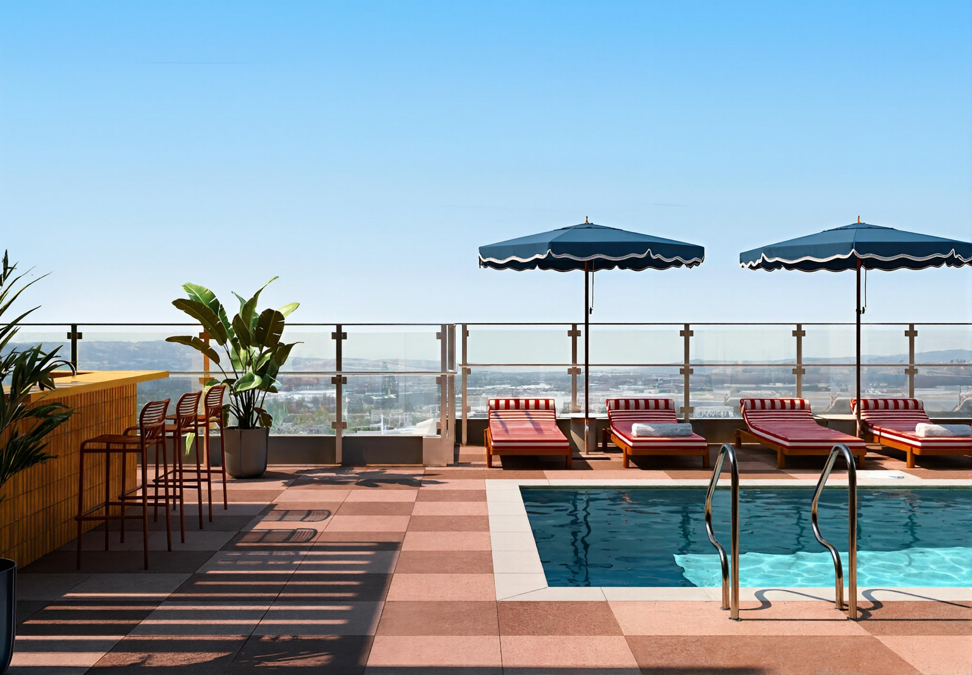 Rooftop pool with chaise lounges, umbrellas, and a bar overlooking a cityscape under a blue sky.