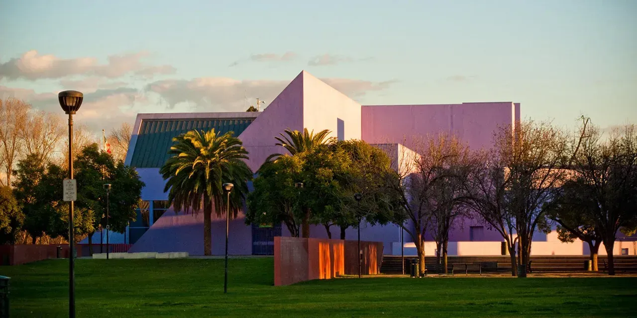 Modern pink building with angular design, green roof, and palm tree in front, set against a sunset sky.