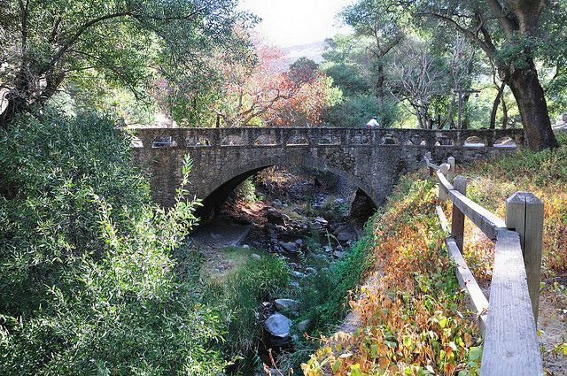 Stone bridge over a creek in a park, surrounded by trees and a wooden fence.