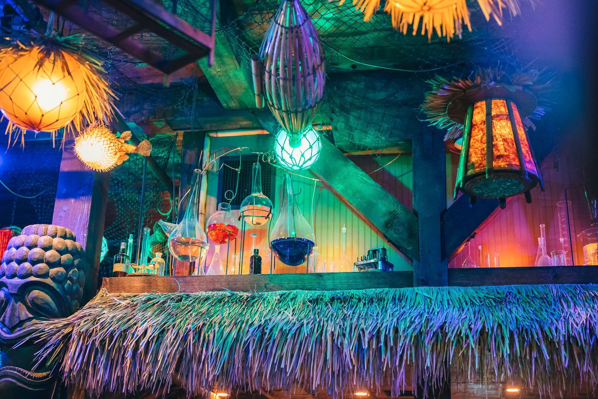 Tiki bar interior with colorful lights and decorations: hanging lanterns, thatched roof, and visible drinks.