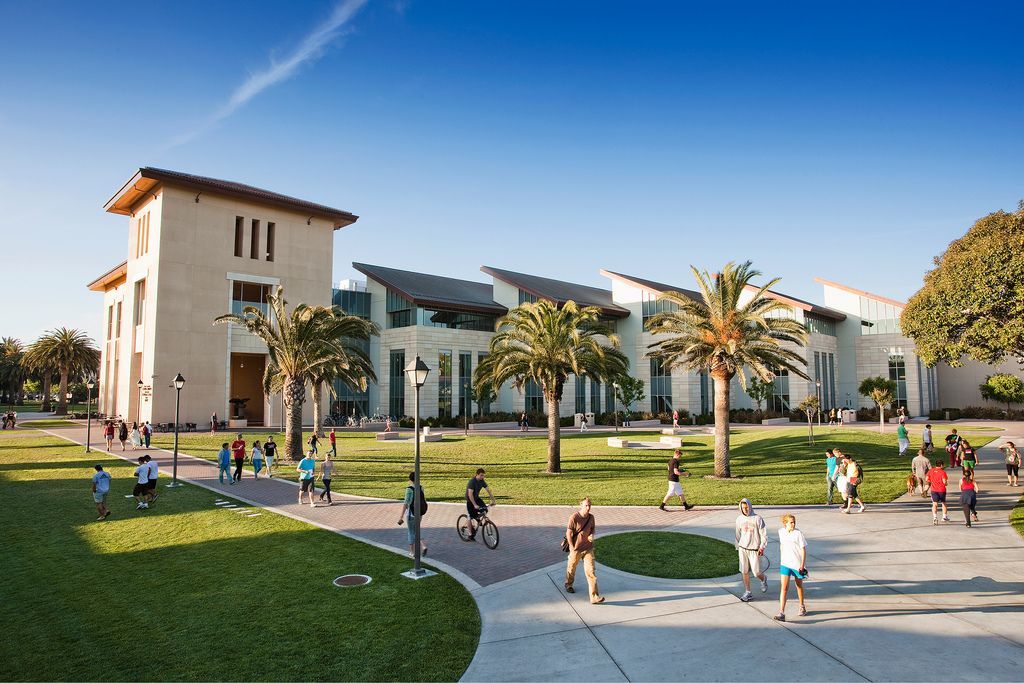 College campus building with people walking on walkways and grassy areas on a sunny day.